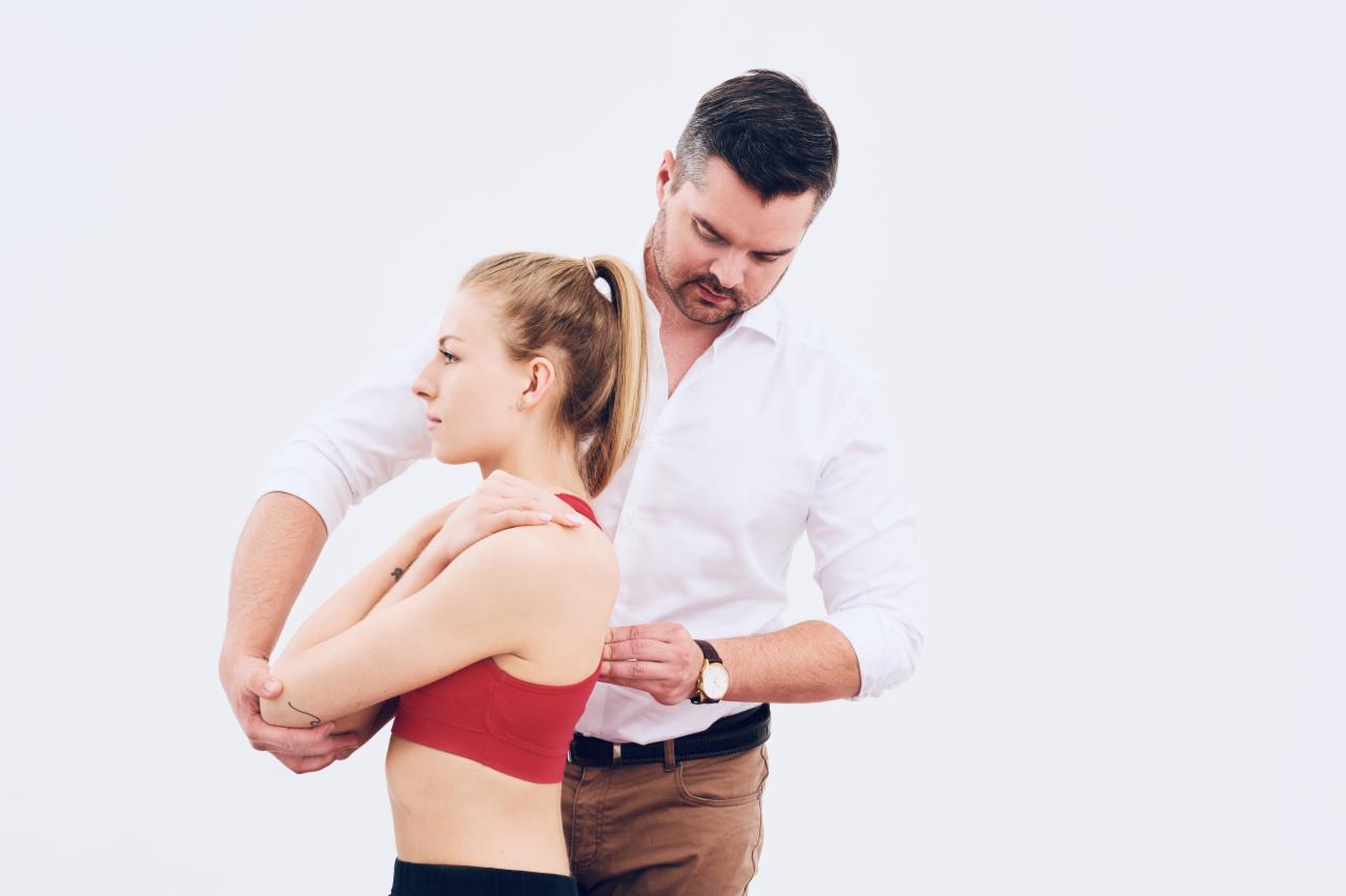 Mężczyzna w białej koszuli i brązowych spodniach mierzy obwód klatki piersiowej kobiety w czerwonym sportowym staniku na białym tle.