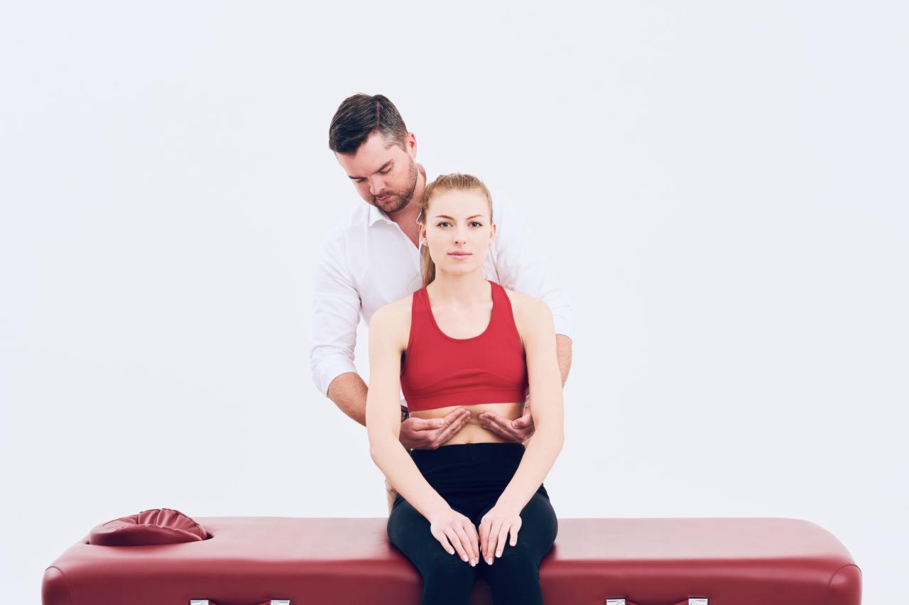 Fizjoterapeuta w białej koszuli bada postawę siedzącej kobiety w czerwonym sportowym topie na bordowym stole do masażu na jasnym tle.