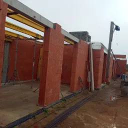 Wznoszenie ścian nośnych z czerwonej ceramiki poryzowanej, wspartych na żółtych belkach stalowych, widoczne rusztowania i zbrojenia, teren budowy w trakcie prac.