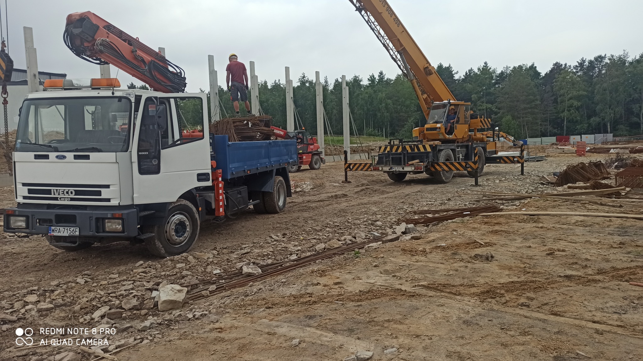 Plac budowy z ciężarówką Iveco Cargo, żurawiem samojezdnym i robotnikiem stojącym na ładunku prętów zbrojeniowych; w tle widoczne słupy konstrukcyjne hali.