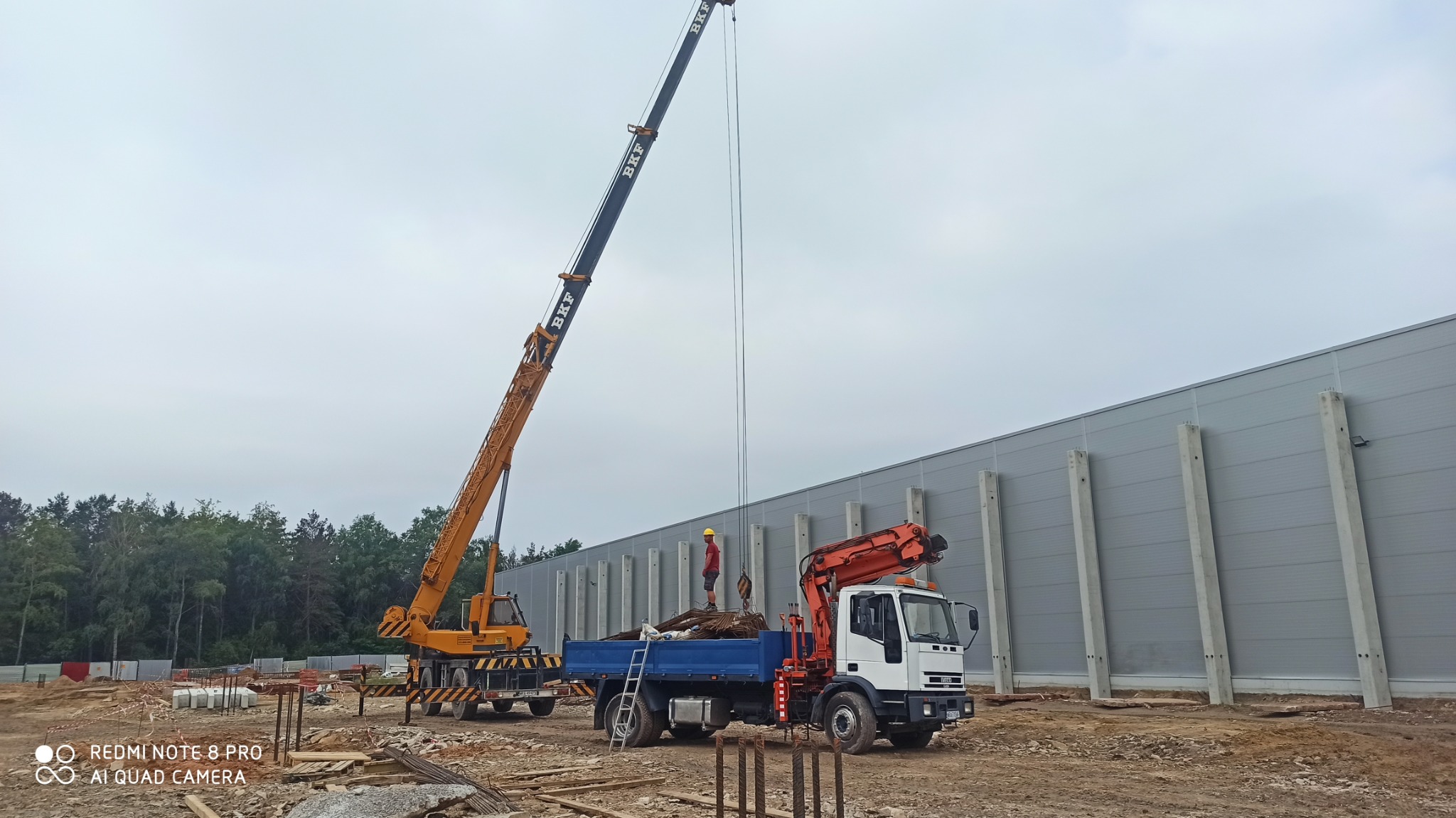 Budowa hali: Żuraw przenosi stalowe elementy na ciężarówkę, pracownik w kasku nadzoruje operację, w tle widoczna szara ściana hali z betonowymi filarami.