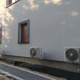 Trzy jednostki zewnętrzne klimatyzacji Rotenso zamontowane na elewacji domu jednorodzinnego, widoczne okna z brązowymi roletami, chodnik z kostki brukowej i przygotowane podłoże pod trawnik.