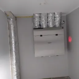 Wnętrze z zamontowaną centralą rekuperacyjną marki reqnet i podłączonymi przewodami wentylacyjnymi, widoczne ślady prac wykończeniowych na podłodze.