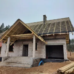 Surowy dom murowany w trakcie budowy dachu, widoczne drewniane elementy konstrukcyjne, komin w trakcie wznoszenia i stos desek na ziemi.