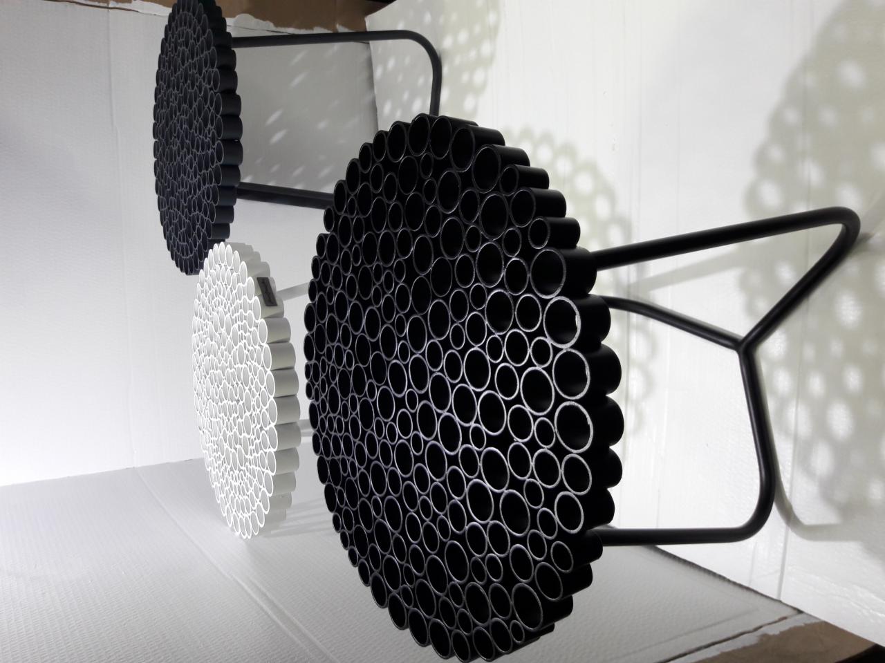 Trzy okrągłe panele dekoracyjne z rurek PCV w kolorze białym i czarnym, zamocowane na metalowych stelażach, tworzące geometryczny wzór na białej ścianie.
