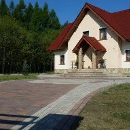 Dom jednorodzinny z czerwonym dachem i podjazdem z kostki w odcieniach brązu i szarości, otoczony zielenią drzew i trawników.