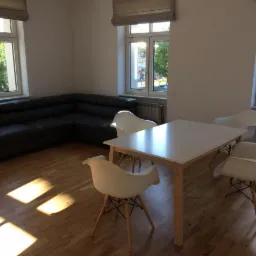 Jasne wnętrze biura z ciemną, skórzaną sofą narożną, białym stołem i czterema białymi krzesłami Eames na drewnianej podłodze, oświetlone promieniami słońca wpadającymi przez okna.