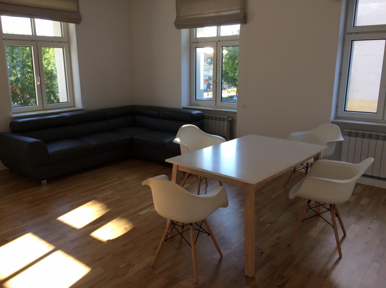 Jasne wnętrze biura z ciemną, skórzaną sofą narożną, białym stołem i czterema białymi krzesłami Eames na drewnianej podłodze, oświetlone promieniami słońca wpadającymi przez okna.