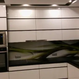 Nowoczesna kuchnia z białymi, lakierowanymi szafkami, czarnym blatem i szklanym panelem ściennym z abstrakcyjnym motywem zieleni i szarości.