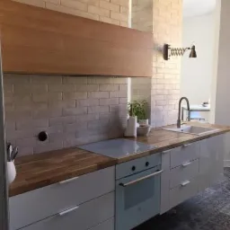 Nowoczesna kuchnia z drewnianym blatem, białymi frontami szafek i ceglaną ścianą, oświetlona lampą w stylu industrialnym.