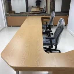 Długi, jasny stół z drewna o nieregularnym kształcie, prawdopodobnie biurowy, z krzesłami w foliowych pokrowcach w tle, widoczne odbicie osoby w lustrze.