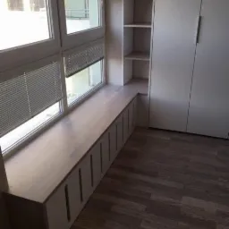 Zabudowa okienna z siedziskiem i szafkami w kolorze jasnego drewna, zintegrowana z wysoką szafą wnękową o białych frontach, na podłodze panele imitujące ciemne drewno.