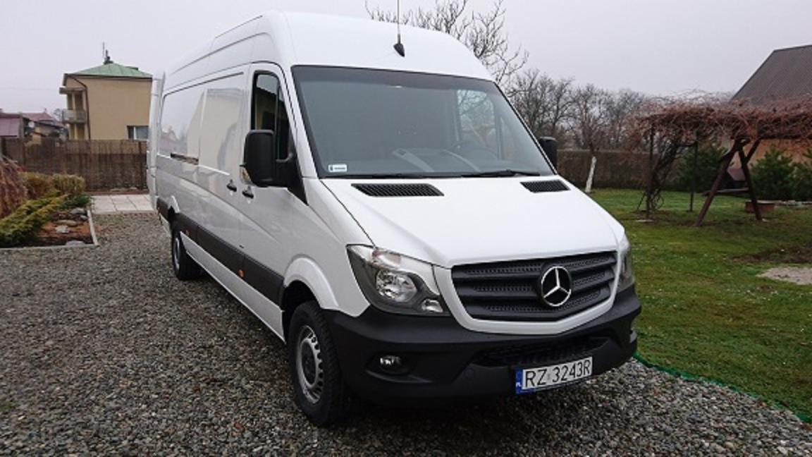 Biały, wysoki furgon Mercedes-Benz Sprinter zaparkowany na żwirowym podjeździe, z widoczną tablicą rejestracyjną RZ 3243R, na tle domu i ogrodu w pochmurny dzień.