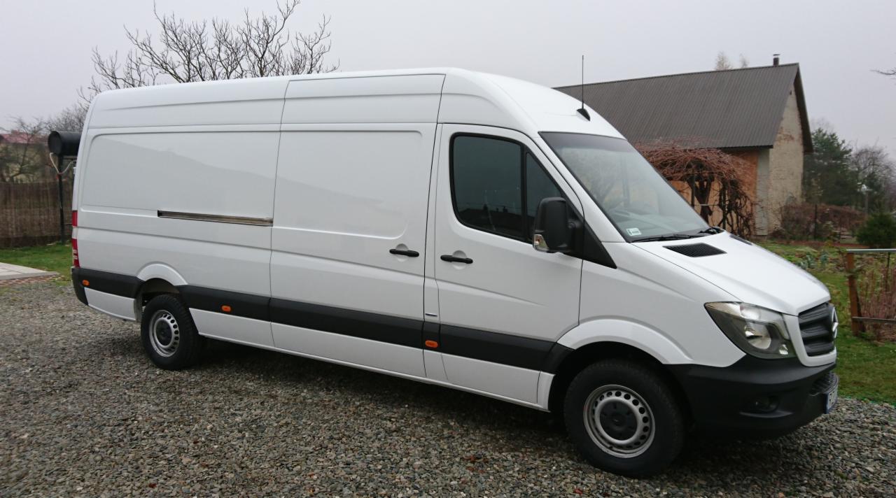 Biały, wysoki furgon Mercedes-Benz Sprinter z przesuwnymi drzwiami bocznymi, zaparkowany na żwirowym podłożu przed budynkiem z szarą dachówką.