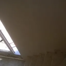 Spojrzenie z góry na fragment poddasza z oknem dachowym i schodami betonowymi w trakcie prac wykończeniowych, widoczne niedoskonałości na ścianie i schodach, drabina oparta o okno.