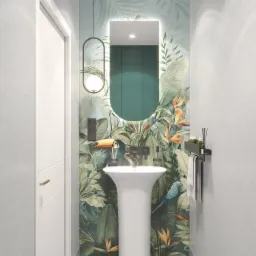 Wąska łazienka z umywalką na postumencie i lustrem z podświetleniem LED, na tle ściany z motywem tropikalnych liści i ptaków. Nowoczesny design, minimalistyczny styl.