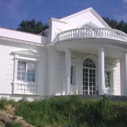 Elegancki, biały dom murowany z kolumnami i balkonem, widoczny z poziomu gruntu, z fragmentem ogrodu w pierwszym planie.