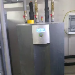 Szara jednostka wewnętrzna z niebieskim wyświetlaczem Alpha Innotec, widoczne rury instalacyjne z zaworami i izolacją, oraz skrzynka elektryczna na ścianie.