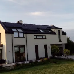 Nowoczesny dom jednorodzinny z panelami fotowoltaicznymi na dachu, widoczne z zewnątrz w porannym świetle, z zadbanym trawnikiem i roślinnością w ogrodzie.