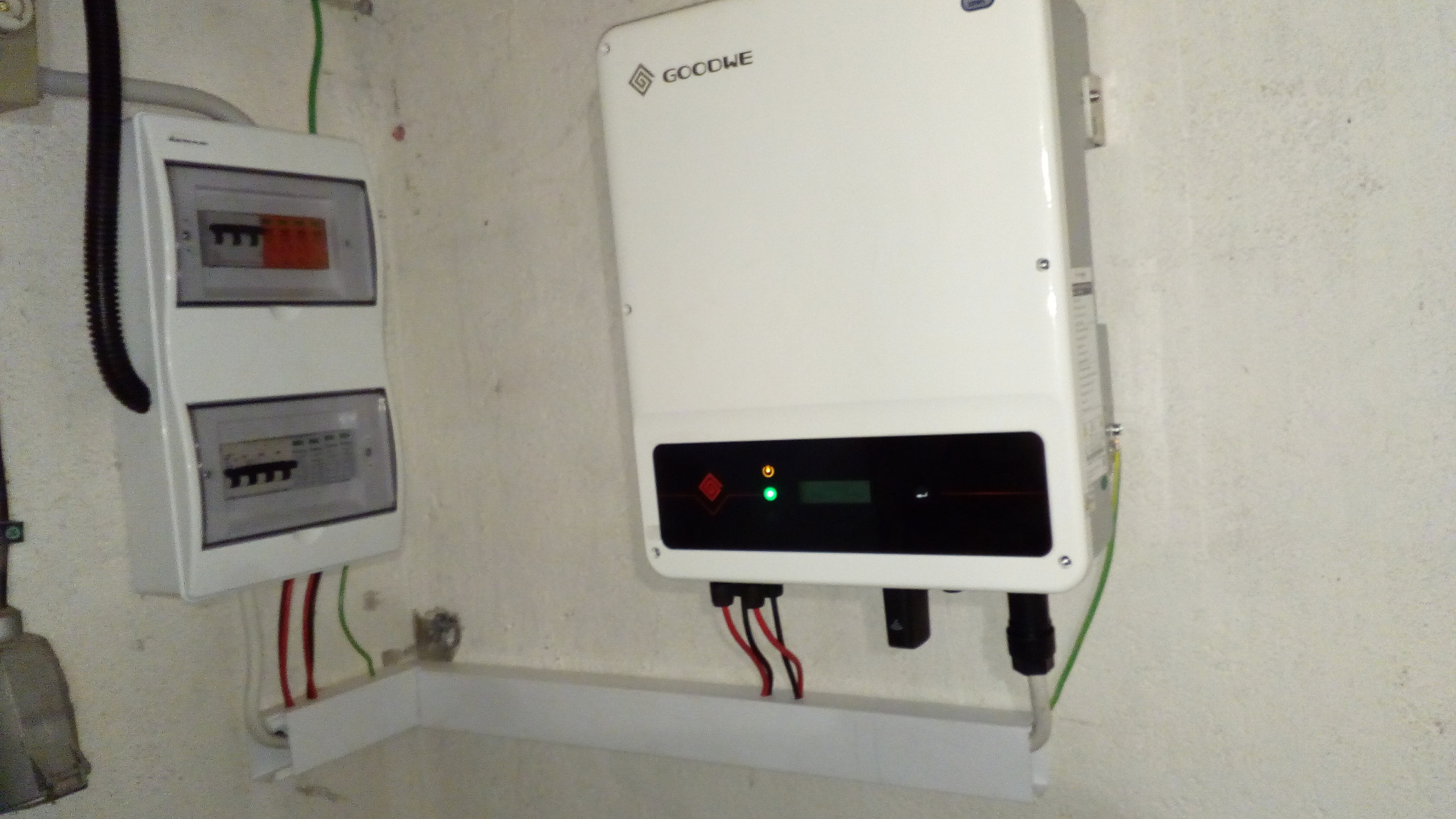 Biała skrzynka elektryczna z bezpiecznikami i biały inwerter z logo Goodwe zamontowane na ścianie wewnątrz budynku, z widocznymi przewodami elektrycznymi i lampkami kontrolnymi.
