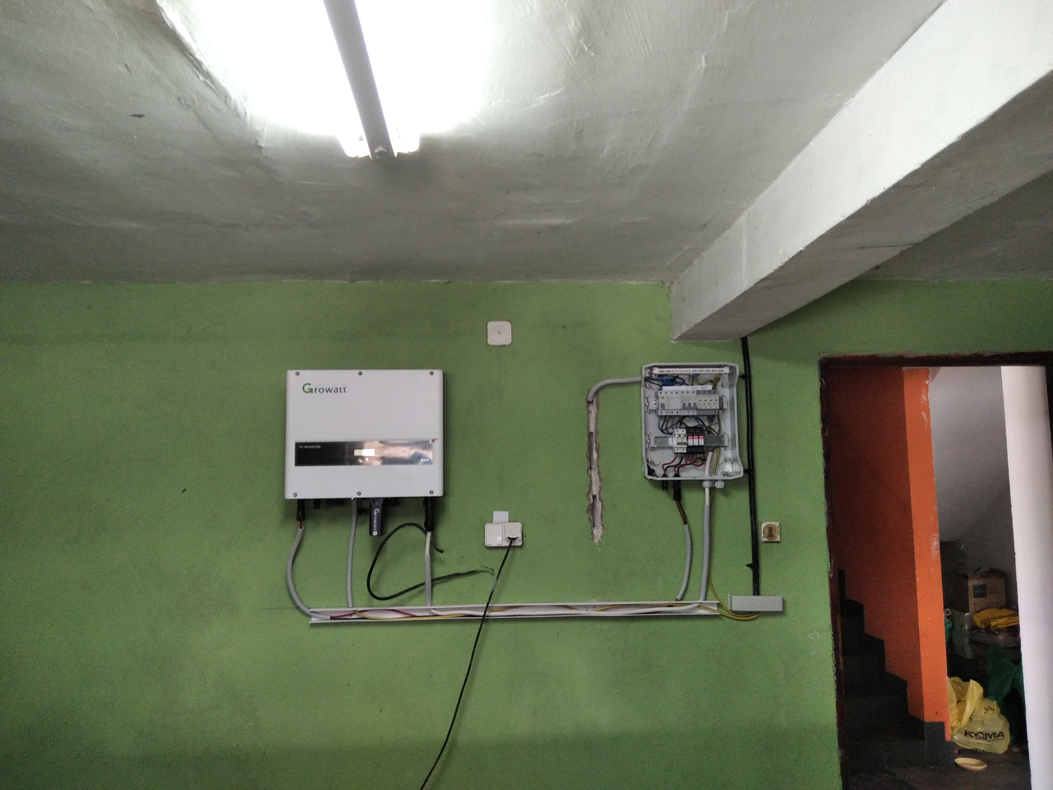 Inwerter fotowoltaiczny marki Growatt i skrzynka elektryczna zamontowane na zielonej ścianie w pomieszczeniu gospodarczym, widoczne przewody w listwie, surowy betonowy sufit z lampą fluorescencyjną.