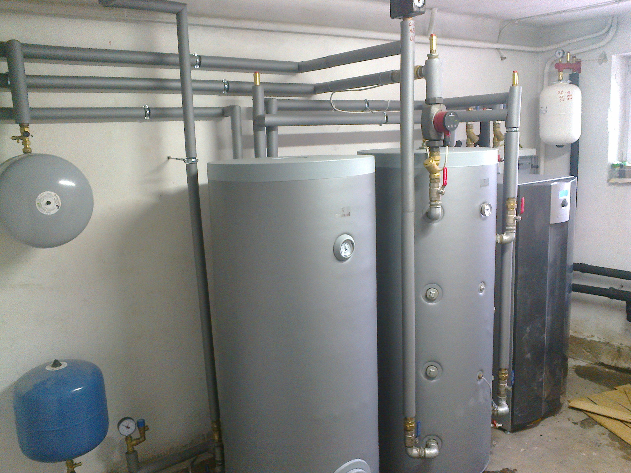 Dwa szare zbiorniki akumulacyjne ciepła w kotłowni, połączone rurami z izolacją termiczną, widoczne również elementy instalacji, takie jak naczynia wzbiorcze i zawory.