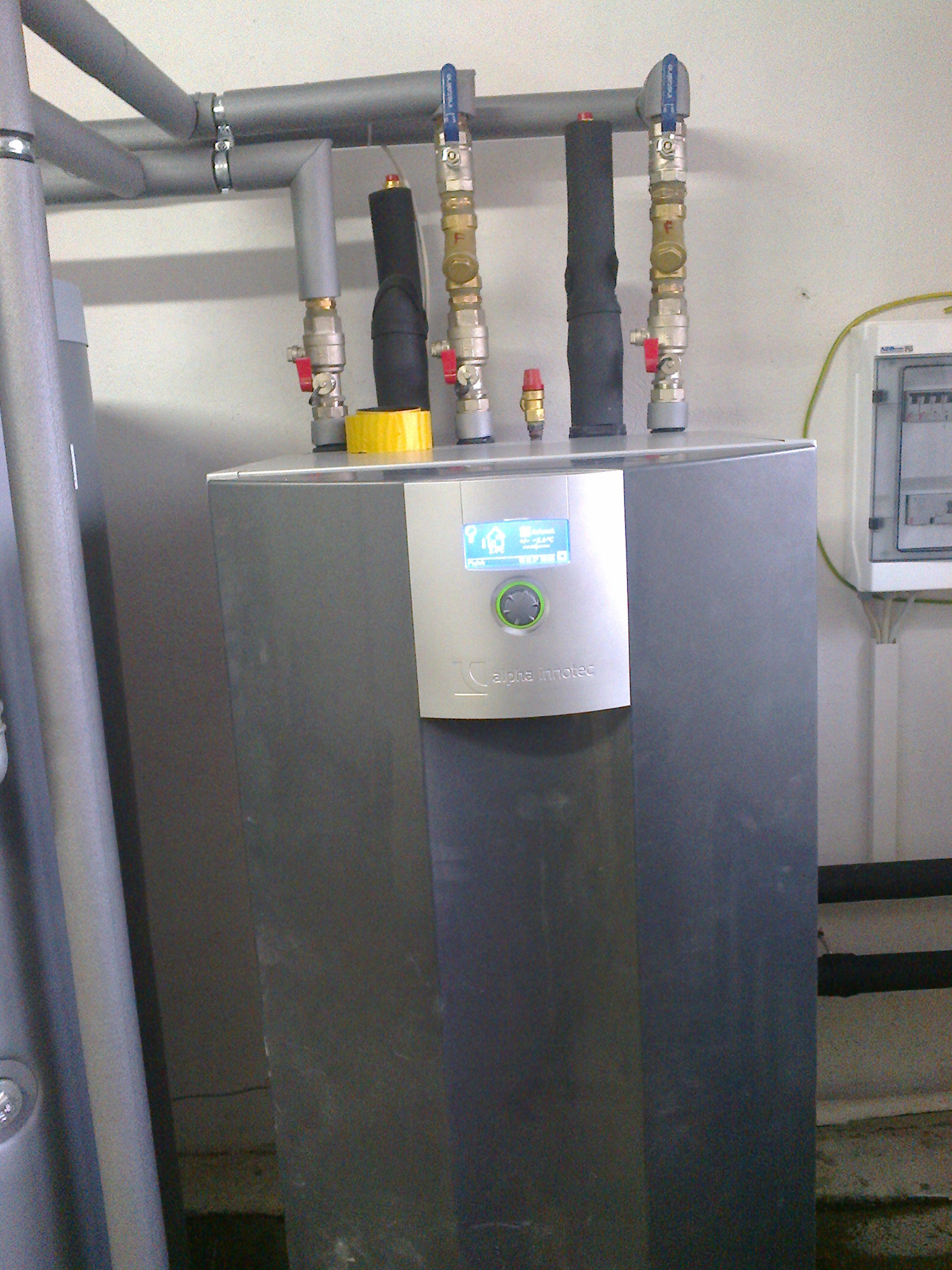 Szara jednostka wewnętrzna z niebieskim wyświetlaczem Alpha Innotec, widoczne rury instalacyjne z zaworami i izolacją, oraz skrzynka elektryczna na ścianie.