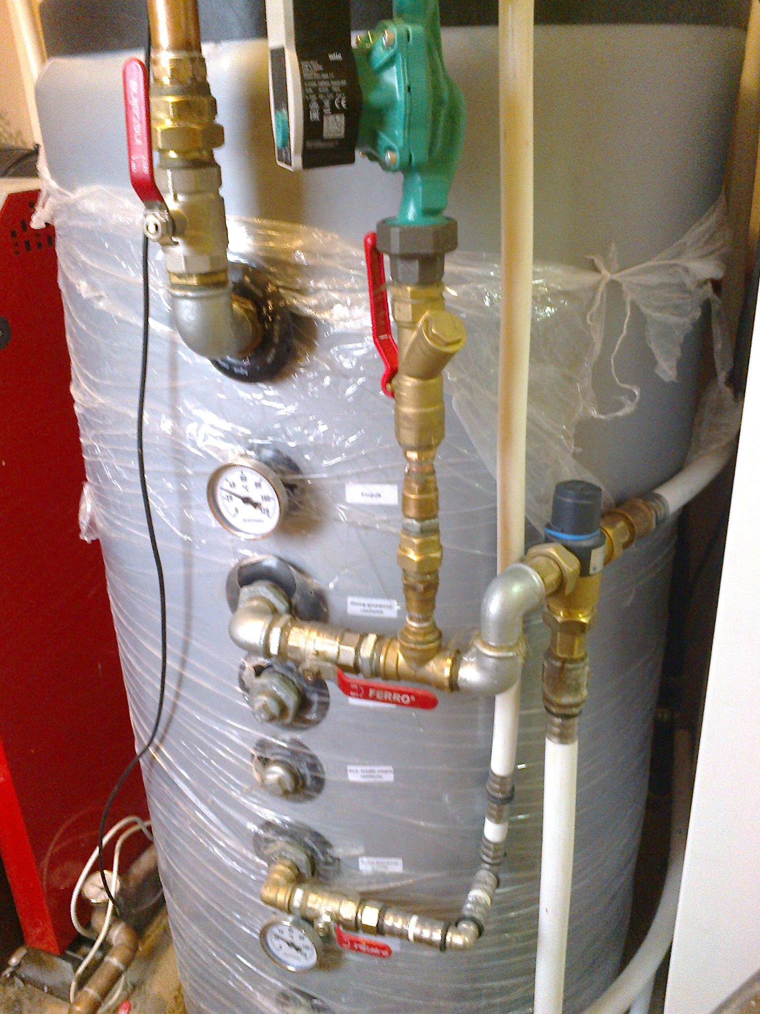 Szary akumulator ciepła owinięty folią ochronną z podłączonymi rurami miedzianymi i zaworami, widoczne manometry i etykiety informacyjne, czerwony korpus kotła w tle.