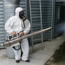 Pracownik w kombinezonie ochronnym i masce gazowej obsługuje zamgławiacz termiczny w pobliżu metalowego silosu zbożowego, z dyszy wydobywa się dym.