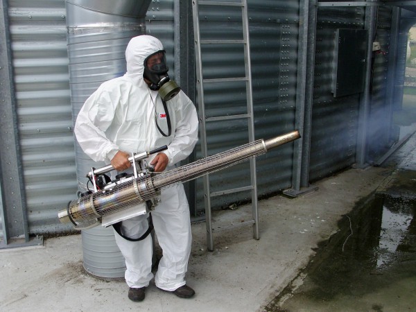 Pracownik w kombinezonie ochronnym i masce gazowej obsługuje zamgławiacz termiczny w pobliżu metalowego silosu zbożowego, z dyszy wydobywa się dym.