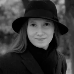 Portret kobiety w czarnym kapeluszu i płaszczu, z delikatnym uśmiechem, na rozmytym tle drzew.