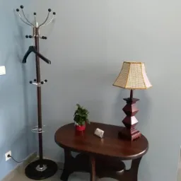 Wnętrze poczekalni z wieszakiem na ubrania, owalnym stolikiem z lampką i małą rośliną doniczkową na tle szarej ściany.