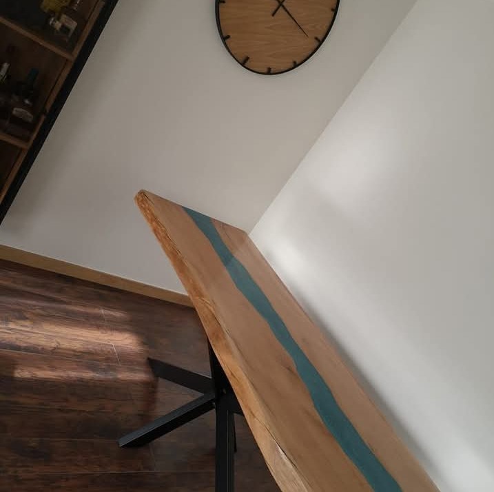 Blat stołu z żywicą epoksydową w kolorze turkusowym, surowe krawędzie drewna, czarne metalowe nogi, drewniana podłoga, zegar na ścianie.