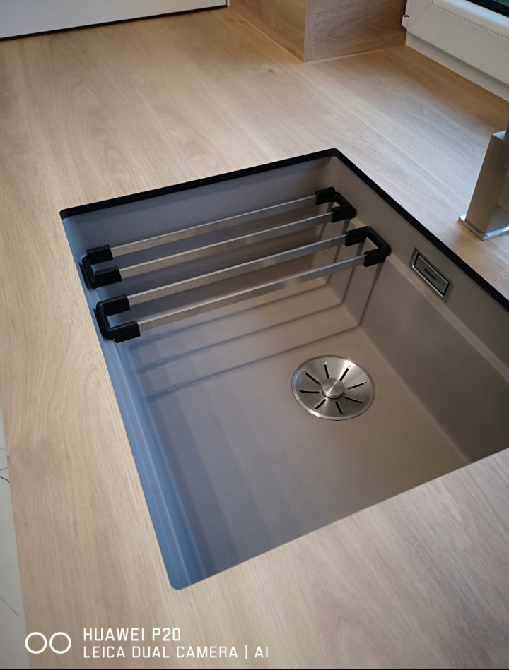 Szary zlew granitowy z ociekaczem, wbudowany w jasny drewniany blat kuchenny, widoczne akcesoria do suszenia naczyń.