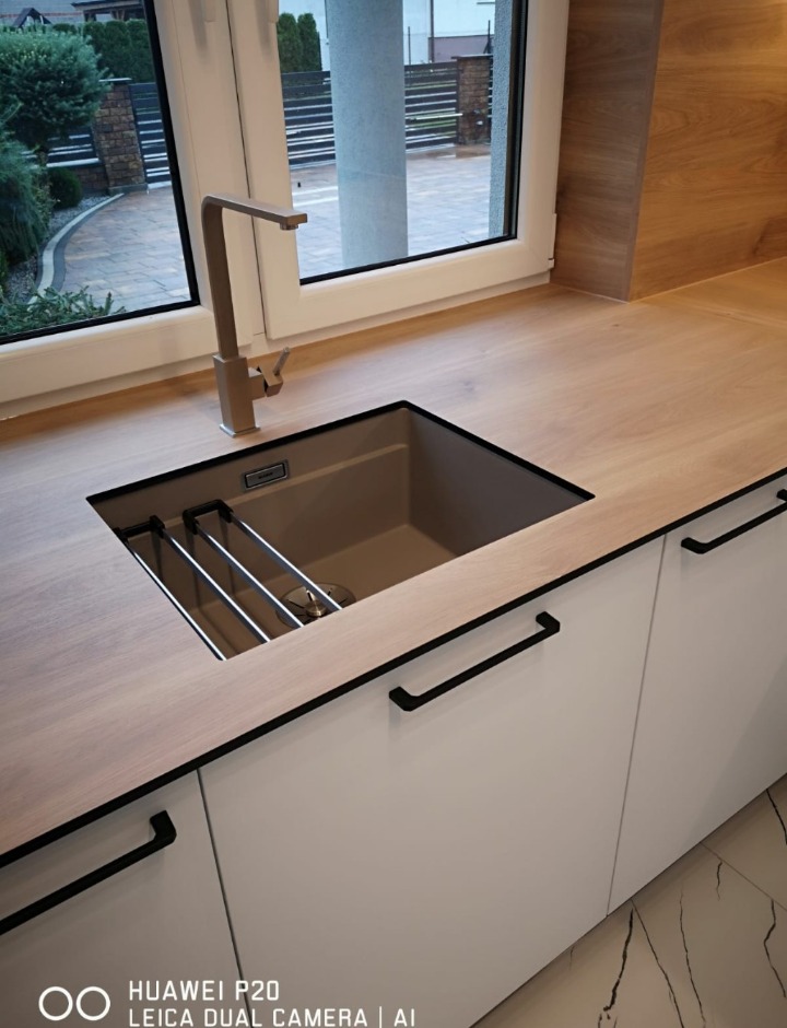 Szczegółowy widok zlewu wpuszczanego w drewniany blat nowoczesnej kuchni z białymi szafkami i czarnymi uchwytami, bateria w kolorze blatu, widok przez okno na zadbany ogród.
