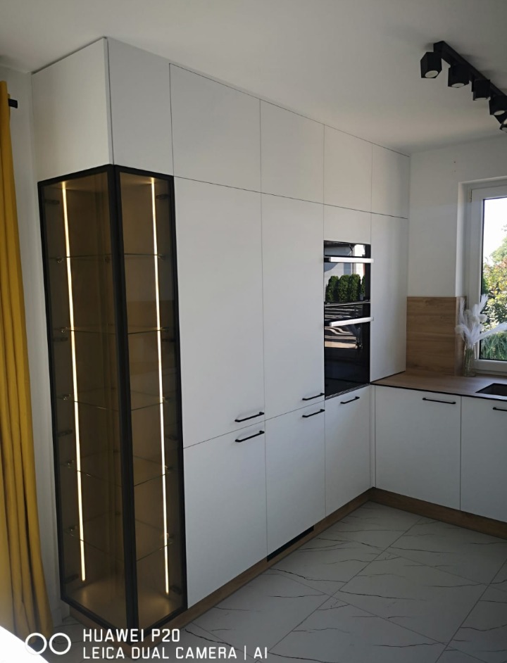 Nowoczesna, biała kuchnia z zabudową sięgającą sufitu, czarnymi uchwytami i przeszkloną witryną z podświetleniem LED, widok na fragment blatu i okno.