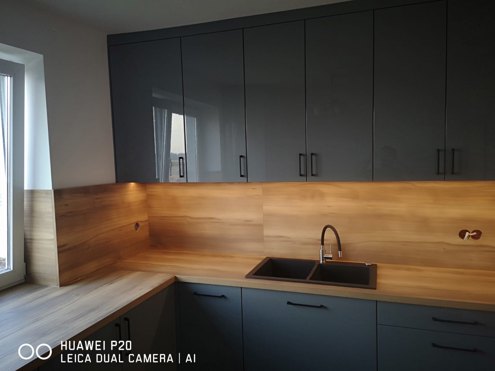 Nowoczesna kuchnia: ciemnoszare, lakierowane szafki wiszące i dolne z drewnianym blatem oraz panelem ściennym, czarny zlewozmywak dwukomorowy z baterią.