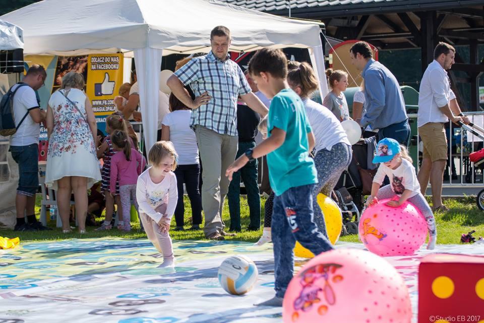 Dzieci bawiące się na zewnątrz podczas festynu: dziewczynka w różowej bluzce i spodniach, chłopiec w turkusowej koszulce, dmuchane piłki, kostka, namiot w tle z napisem 'Dołącz do nas na Facebooku'.