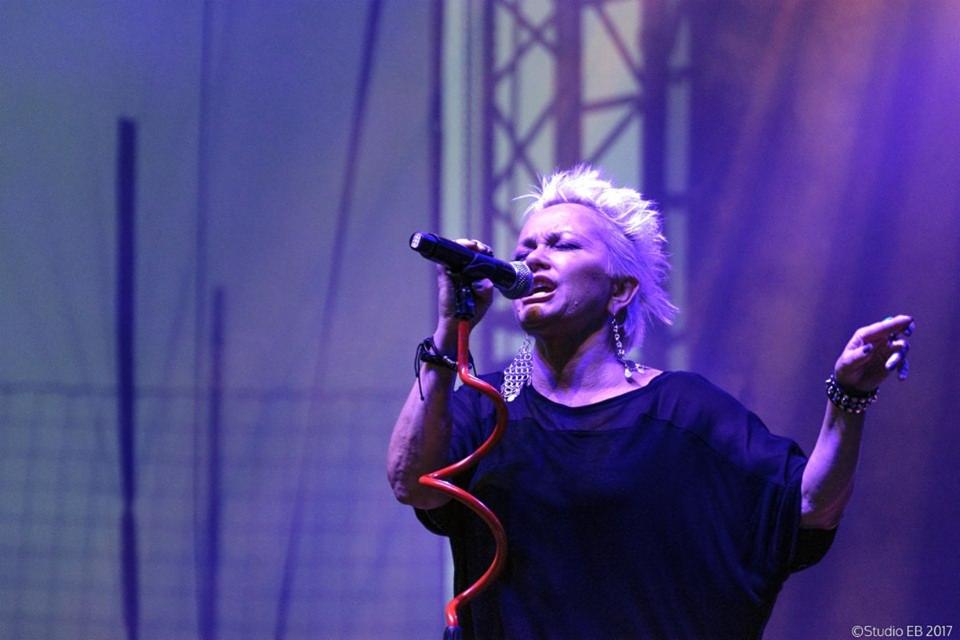 Piosenkarka z krótkimi blond włosami, w czarnej bluzce, śpiewa do mikrofonu na scenie oświetlonej niebieskim światłem, z czerwonym kablem mikrofonu w kształcie spirali.