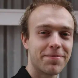 Portret młodego mężczyzny z lekkim uśmiechem, jasna karnacja, krótko ostrzyżone włosy, na tle białej ściany z pionowymi szarymi paskami.