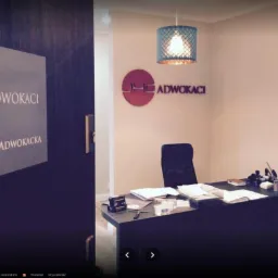 Wnętrze kancelarii adwokackiej z logo JHC Adwokaci na ścianie i drzwiach, biurko z dokumentami i lampy wiszące.