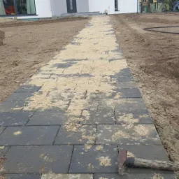 Układanie ciemnoszarej kostki brukowej na piasku, widoczny młotek, przygotowane podłoże i fragment domu w tle.