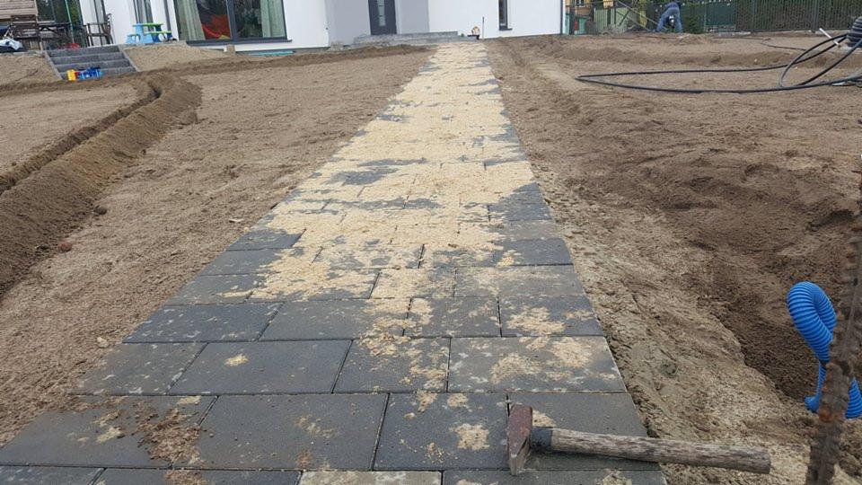 Układanie ciemnoszarej kostki brukowej na piasku, widoczny młotek, przygotowane podłoże i fragment domu w tle.