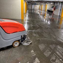 Steamlux - Szorowarka automatyczna w trakcie pracy na mokrej posadzce garażu podziemnego. Widoczne ślady po czyszczeniu, żółte filary i instalacje pod sufitem.