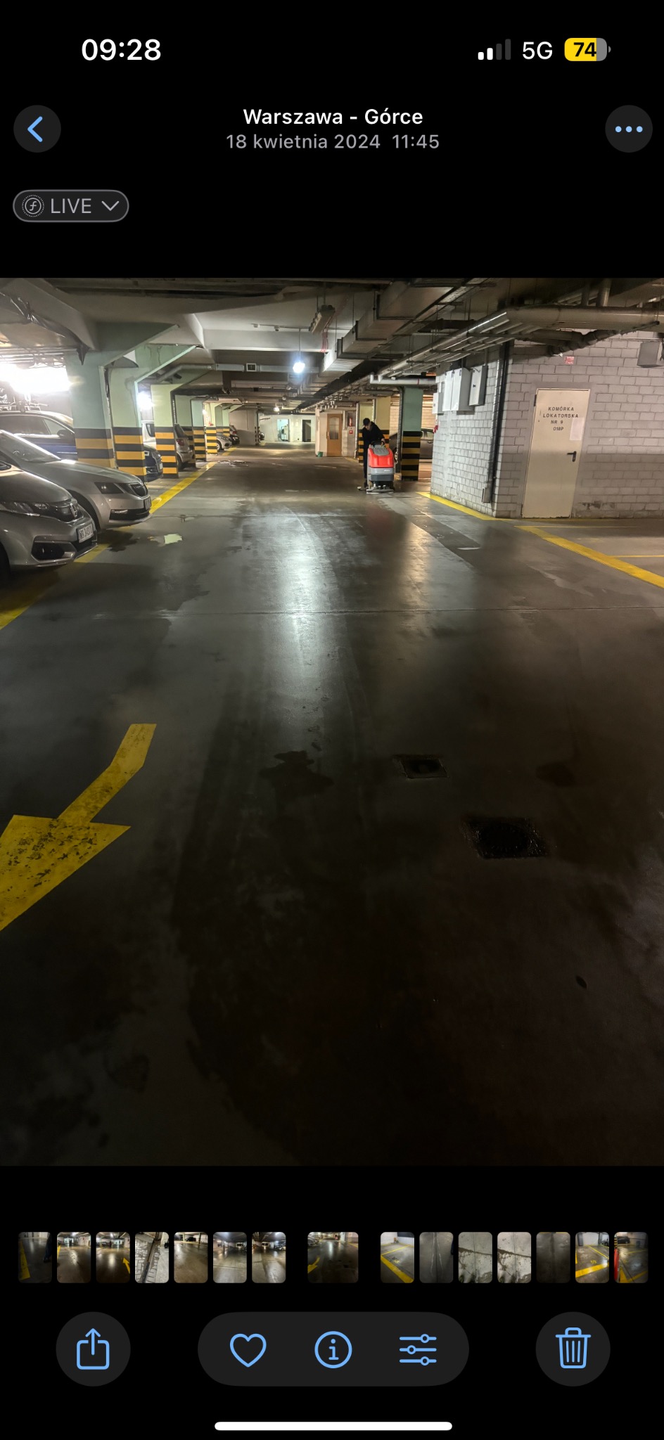 Pracownik z maszyną czyszczącą posadzkę w garażu podziemnym. Widoczne samochody i żółte oznakowania na podłodze. Oświetlenie sufitowe.