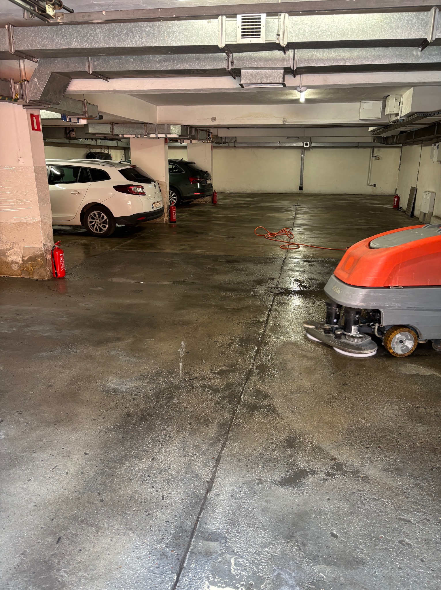 Czyszczenie posadzki w garażu podziemnym za pomocą profesjonalnej maszyny czyszczącej. Widoczne samochody i hydranty.