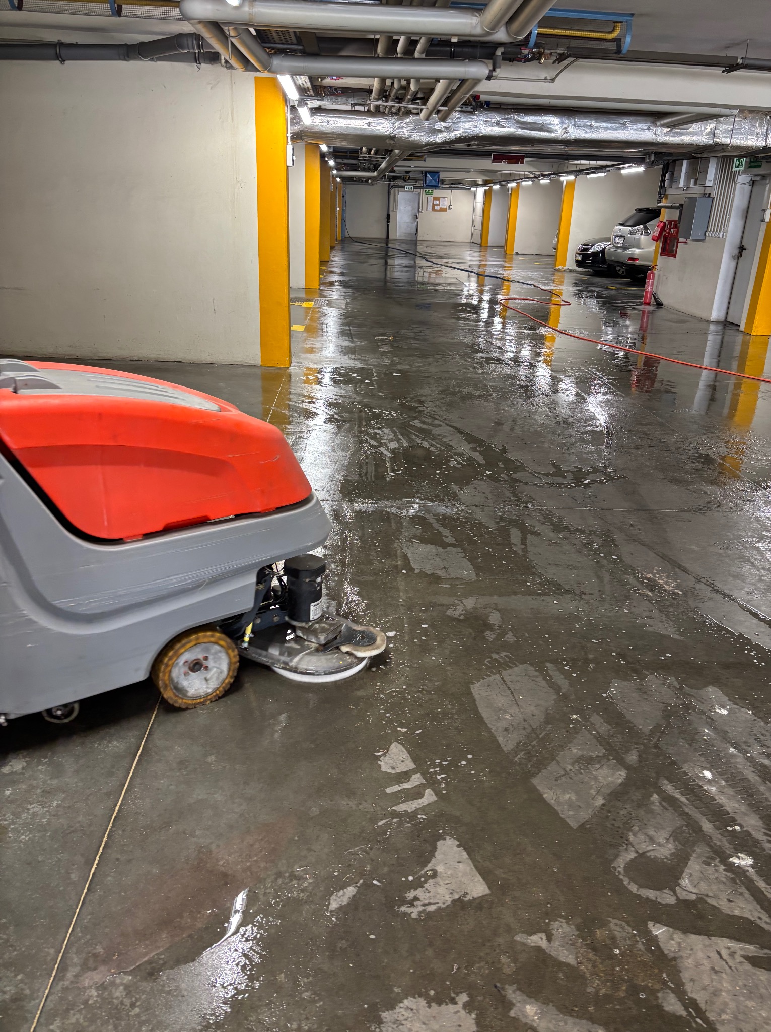 Szorowarka automatyczna w trakcie pracy na mokrej posadzce garażu podziemnego. Widoczne ślady po czyszczeniu, żółte filary i instalacje pod sufitem.