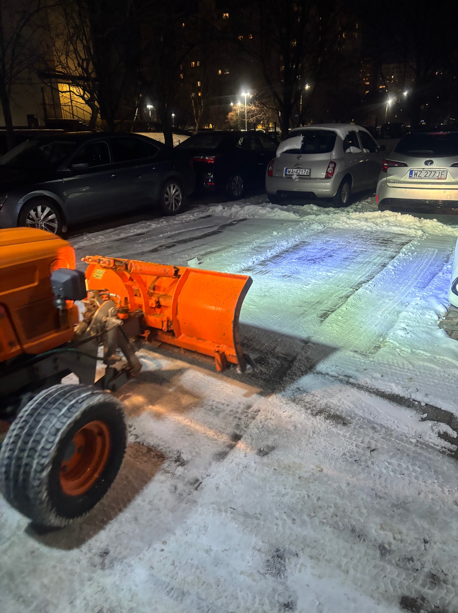 Pomarańczowy pług śnieżny odśnieża parking samochodowy nocą. Widoczne ślady opon na śniegu i zaparkowane auta w tle.