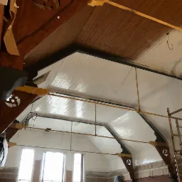 Wnętrze budynku w Jurgowie z drewnianą konstrukcją dachu w trakcie malowania na biało. Widoczne elementy dekoracyjne i rusztowanie.