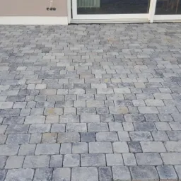 Ułożona szara kostka brukowa na podjeździe przed domem, widoczne przesuwne drzwi tarasowe i fragment elewacji.